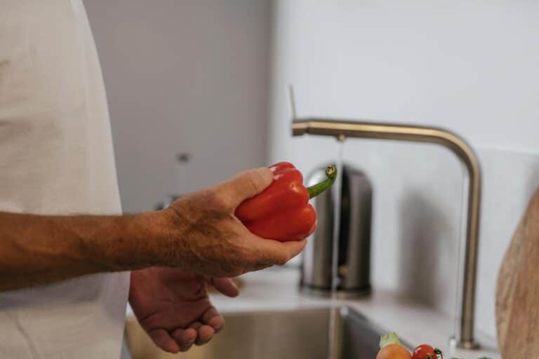 A person's hand holding a red bell pepper while washing it under a kitchen tap.